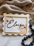 Custom floral baby name sign with scalloped white frame, featuring the name Eloise Lynn in 3D script and printed text with soft blush florals.