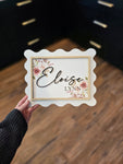 Personalized baby girl name sign displayed in a nursery, with floral details and a scalloped edge frame, perfect for nursery wall decor.