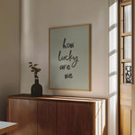 Sage green typography art print reading "how lucky are we," framed and styled in a modern sunlit room with minimalist decor.