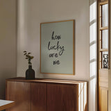Sage green typography art print reading "how lucky are we," framed and styled in a modern sunlit room with minimalist decor.