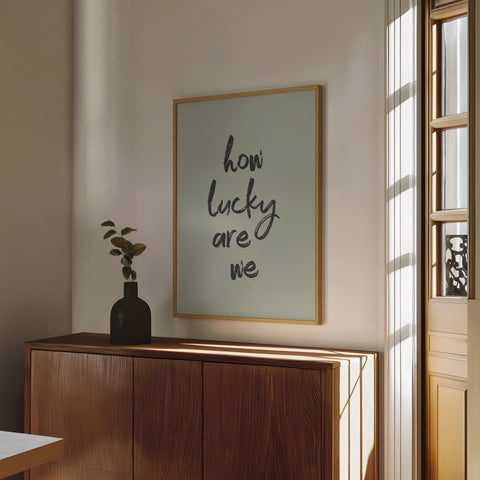 Sage green typography art print reading "how lucky are we," framed and styled in a modern sunlit room with minimalist decor.